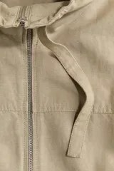 Campera beige en mezcla de lino, con cuello elevado con cordón ajustable, cierre frontal y bolsillos laterales con solapa y botón. Presenta hombros caídos y elástico revestido en el bajo.