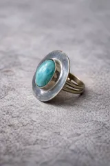 Anillo ajustable de alpaca con una piedra central ovalada de color turquesa. El engaste presenta un bisel circular amplio y texturizado que rodea la piedra. La banda del anillo es doble o triple.
