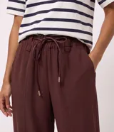 Pantalón carrot marrón de tiro alto, confeccionado en mezcla de viscosa y poliéster, con cintura elástica fruncida, cordón de ajuste tipo lazo con punteras metálicas, bolsillos y recortes estructurados en las piernas.