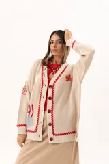 Saco tipo cardigan oversize de punto color beige, con diseño temático marino que incluye bordados de corales, peces, estrellas de mar y tortugas. Presenta detalles de galón picot en color rojo en los bordes y puños, cierre frontal con botones al tono y corte holgado.