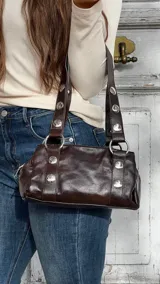 Cartera de hombro color marrón oscuro, confeccionada en material con acabado brillante. Presenta un diseño estructurado con detalles de remaches metálicos plateados en las correas y en el cuerpo de la cartera, además de argollas metálicas que conectan las correas con la base.
