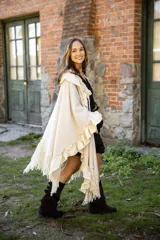 Ruana de tejido liviano color beige, con diseño abierto y detalles de volados en mezcla de lino y algodón. Presenta terminaciones con flecos en el ruedo y cordones decorativos con borlas en el frente.