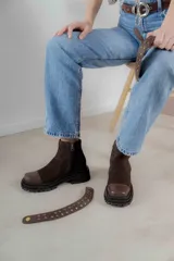 Bota corta de gamuza marrón con puntera de cuero liso, suela track negra y correa desmontable con tachas metálicas.