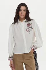 Blusa de mezcla de algodón y lino color blanco, con mangas abullonadas y cierre frontal de botones. Presenta detalles de bordado geométrico a cuadros en el cuello y los puños, y un gran bordado floral en el pecho.