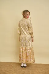 Vestido largo dorado con estampado floral y detalles metalizados, escote en V, mangas largas con puños ajustables con cordones y volados en la falda.