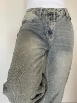 Pantalón de jean rígido de corte recto, con acabado lavado y aplicación de brillos distribuidos por toda la prenda. Presenta diseño de cinco bolsillos, cierre frontal con botón y tiro medio.