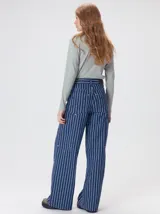 Pantalón de tiro medio y pierna ancha confeccionado en denim de algodón grueso. Presenta un diseño a rayas verticales en tonos azul y celeste, con costuras en contraste color rojo, bolsillos laterales tipo cargo y remaches metálicos.