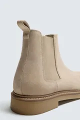 Bota estilo Chelsea de hombre, confeccionada en piel con acabado de serraje en color beige. Presenta elásticos laterales al tono, horma redonda y suela de goma a contraste con ligero tacón.