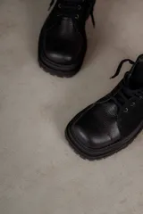Borcegos de punta cuadrada con diseño acordonado, combinando paneles de cuero texturizado y gamuza en tonos marrones. Presentan una suela track gruesa de goma negra y cordones negros.