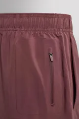Pantalones cortos deportivos color marrón rojizo, confeccionados en tejido técnico ligero y elástico. Presentan cintura elástica ajustable con cordón interior, bolsillos frontales y un bolsillo trasero con cierre de cremallera. Los laterales tienen perforaciones para transpirabilidad y aberturas laterales en el bajo.