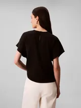 Blusa Calvin Klein de manga corta en color negro, confeccionada en tejido fluido de crêpe de chine. Cuenta con un diseño de cuello redondo con escote en V y una barra metálica decorativa en el centro del cuello.