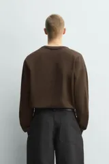 Camiseta de manga larga y corte relajado en color marrón oscuro. Presenta un diseño de doble capa en el cuello y el ruedo, con un borde interior en color blanco que crea un efecto de contraste.