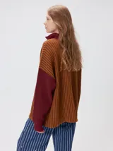 Pullover de tejido acanalado grueso con cuello alto y cierre frontal parcial. Presenta un diseño de bloques de color, con cuerpo en tono beige y mangas en contraste con rayas verticales en tonos rojizos. Incluye un detalle bordado en el pecho.