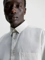 Camisa blanca de manga larga con cuello clásico, cierre frontal con botones, bolsillo de parche en el pecho y logo CK bordado en el bolsillo.