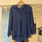 Camisa de lino color azul jean, con cuello camisero, cierre frontal de botones, bolsillo en el pecho y diseño de bajo asimétrico, más largo en la parte trasera.