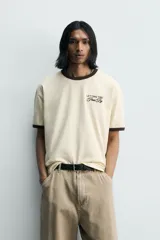 Camiseta de corte regular, color beige con cuello y bordes de mangas en color marrón oscuro. Presenta un diseño de texto bordado en el pecho con la frase 'Letting Time Pass By'.