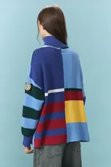 Sweater de tejido de punto grueso con diseño de bloques de color y rayas horizontales en tonos azul, rojo, amarillo y verde. Presenta cuello alto con cierre frontal parcial y mangas largas con puños ajustados.