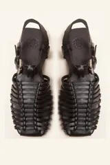 Sandalia de cuero negro estilo huarache, con puntera cerrada mediante tiras finas entretejidas a mano, tira ajustable al tobillo con hebilla metálica y suela plana con taco de 1 cm.