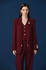 Saco de mujer color bordó con solapas redondeadas, cierre frontal de un botón dorado y bolsillos laterales con solapa. Presenta un parche decorativo en la manga izquierda y un broche metálico en la solapa.