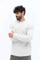 Remera de manga larga color blanco roto o gris muy claro, con escote redondo y un pequeño logo circular estampado en el pecho.