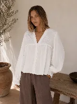 Camisa blanca de corte amplio con escote en V y cierre frontal de botones. Presenta detalles de bordado floral en el canesú y mangas largas abullonadas con puños elásticos con volados.