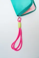 Billetera rectangular con cierre, color celeste pálido, con parche frontal negro que lleva las letras "CNJ" en relieve. Incluye un llavero de muñequera con cordón trenzado en rosa fuerte y verde lima.