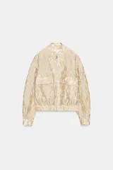 Chaqueta tipo bomber de cuello redondo y manga larga, confeccionada en tejido jacquard con diseño floral en tonos dorados y crema. Presenta cierre frontal con cremallera, bolsillos delanteros de plastrón con solapa y puños y bajo elásticos.