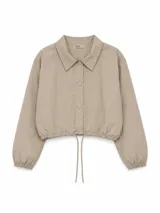 Campera corta de tela técnica con acabado ligeramente arrugado, color beige. Presenta cuello camisero, cierre frontal con botones a presión, mangas largas con puños elásticos y ruedo ajustable con cordón.