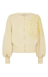 Cardigan de punto color amarillo claro, con cierre frontal de botones metálicos, cuello redondo y mangas largas abullonadas con puños ajustados. Presenta un diseño asimétrico con un panel de punto calado texturizado aplicado sobre uno de los hombros.