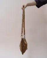 Bolso de hombro tipo hobo confeccionado en gamuza suave color marrón oscuro. Presenta un diseño de media luna con bordes trenzados, cierre superior con tiradores de cuero anudado y una correa ajustable y desmontable con herrajes metálicos.