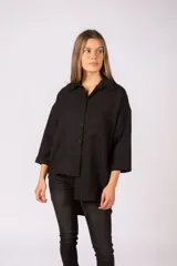 Camisa negra de corte oversize, con cuello clásico, cierre frontal de botones y bolsillo en el pecho. Presenta mangas tres cuartos y un diseño con ruedo irregular, más largo en la parte posterior.
