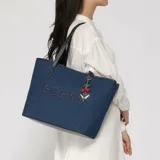 Cartera tipo tote bag Guess modelo Keira Denim, color azul con acabado de mezclilla. Cuenta con asas dobles de cuero sintético azul oscuro, logo "GUESS" bordado en relieve al frente y un llavero colgante con dijes de cerezas y forma triangular metálica.