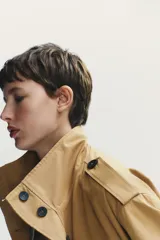 Chaqueta trench corta color beige, con cuello subido, manga larga con trabillas en hombros y mangas, solapas en pecho, bolsillos con solapa en delantero, cinturón lazada en mismo tejido, forro interior y cierre frontal con botones ocultos por solapa.