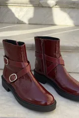 Bota corta de cuero charolado color bordó, con suela track negra, cierre lateral y tiras decorativas con hebillas metálicas plateadas.