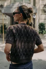 Chaqueta de punto texturizado con cuello redondo y manga corta. Presenta un patrón de rombos en tonos marrón oscuro, negro y blanco. Cierre frontal con botones oscuros.