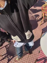 Campera tejida con textura vanizada a rayas horizontales en blanco y negro, con cierre frontal y flecos negros en forma de V en el pecho.