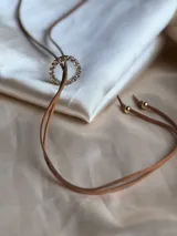 Choker marrón de gamuza con dije de doble aro entrelazado, uno liso y otro con incrustaciones de strass. Los extremos del choker tienen detalles de cuentas doradas.