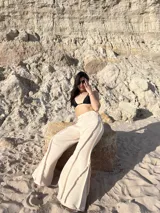 Pantalón largo color beige claro, de tejido ligero con textura, tiro alto y cintura elástica. Presenta detalles deshilachados o con terminación cruda en los bordes verticales de la pierna, creando un efecto drapeado y fluido.