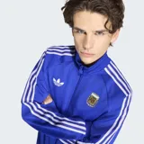 Campera deportiva Adidas de la Selección Argentina, color azul con las icónicas tres tiras blancas en las mangas. Presenta cuello alto, cierre frontal, logo del Trifolio bordado en el pecho y escudo de la AFA.