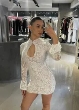 Vestido corto de encaje blanco con forro en tono nude, diseño ajustado, cuello alto con abertura frontal tipo gota y mangas largas con terminación acampanada.