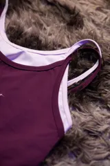 Top deportivo ajustado color morado oscuro con doble capa de tela y tirantes finos blancos. Presenta un logo manuscrito en tono rosado claro en el pecho.