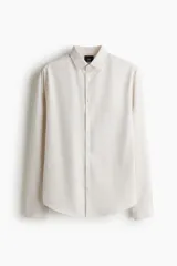 Camisa de vestir color beige claro, de manga larga, con cuello inglés y cierre de botones. Presenta un corte slim fit y bajo ligeramente redondeado.