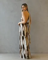 Vestido largo de satén con estampado abstracto en tonos tierra, escote halter con cuello alto y espalda descubierta. Presenta un diseño drapeado en la cintura y una abertura lateral profunda en la falda.