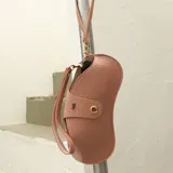 Estuche para lentes de diseño ergonómico y minimalista, confeccionado en cuero sintético con textura granulada. Cuenta con cierre magnético frontal, costuras a la vista en color contrastante y una correa de mano extraíble.