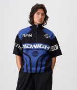 Remera de tricot liviano Blue Steel con corte boxy, cuello polo alto y cierre frontal parcial de un cuarto. Presenta diseño jacquard integral con gráficos y lettering en tonos azul, negro y blanco, mangas cortas y estilo de inspiración deportiva.