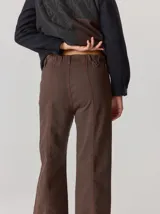 Pantalón de corte relajado color marrón, confeccionado en una mezcla de algodón y elastano. Presenta costuras en contraste color rojo en bolsillos y bragueta, con cierre de botón metálico.