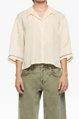 Camisa corta de corte boxy, confeccionada en tejido fantasía texturado color crema o amarillo muy pálido. Presenta cuello camisero, botones frontales y manga 3/4 con sisa ajustable mediante tiras de tejido, además de pespuntes decorativos negros que realzan el borde de las mangas.