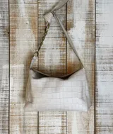 Cartera de hombro color beige con textura tipo cocodrilo, confeccionada en gamuza. Presenta un diseño estructurado con una cadena metálica decorativa en la parte superior del asa y cierre principal.