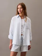 Camisa celeste de gasa doble, con cuello clásico, cierre frontal con botones, bolsillo de parche en el pecho y mangas largas con solapa enrollable.