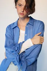 Camisa de cuello solapa y manga larga con detalle de lazos en los puños. Presenta un diseño de rayas verticales a tono y cierre frontal con botones ocultos.